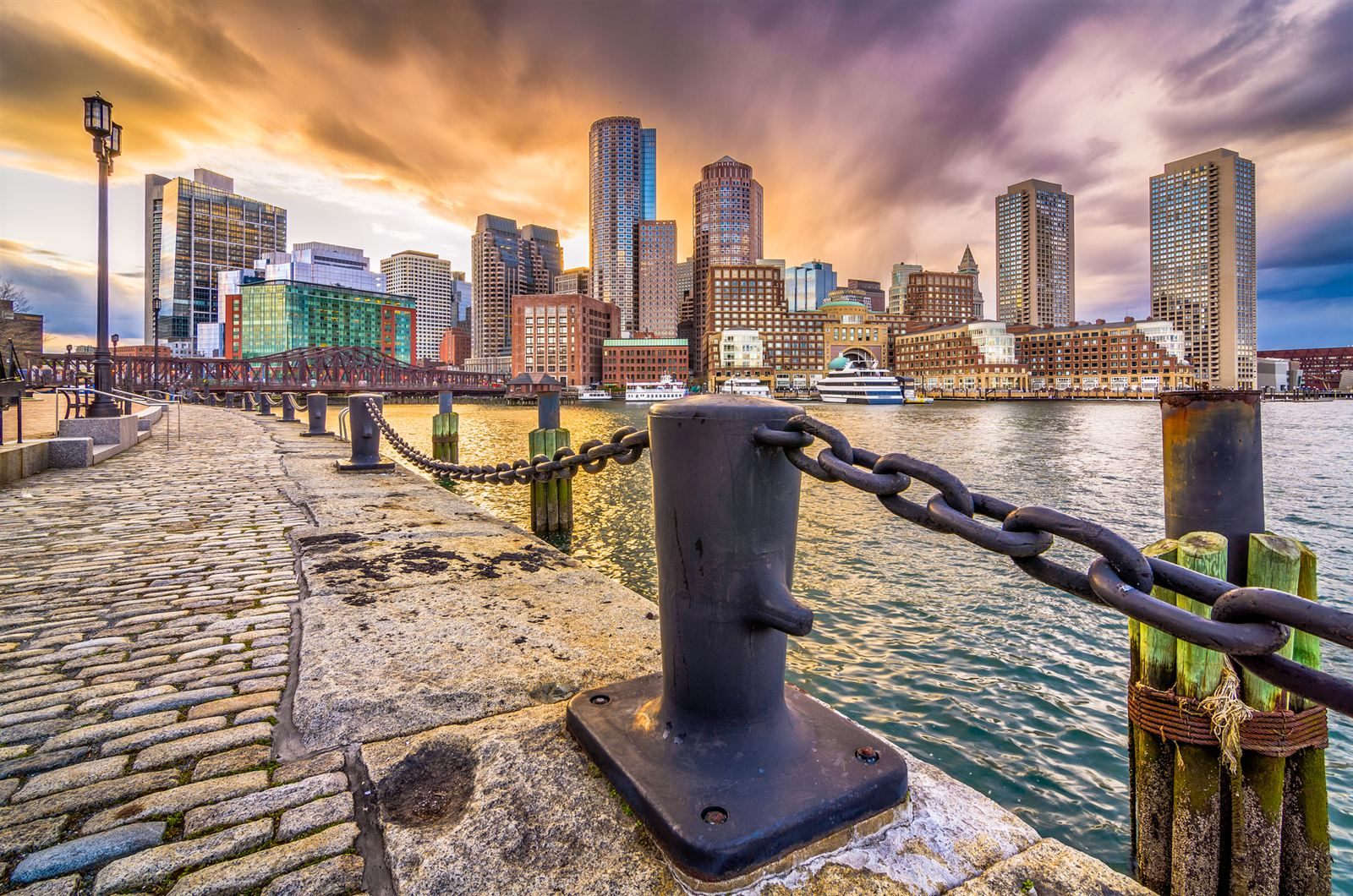 Boston skyline across the harbor at sunset, seen from a stone wharf with an iron bollard and chain
