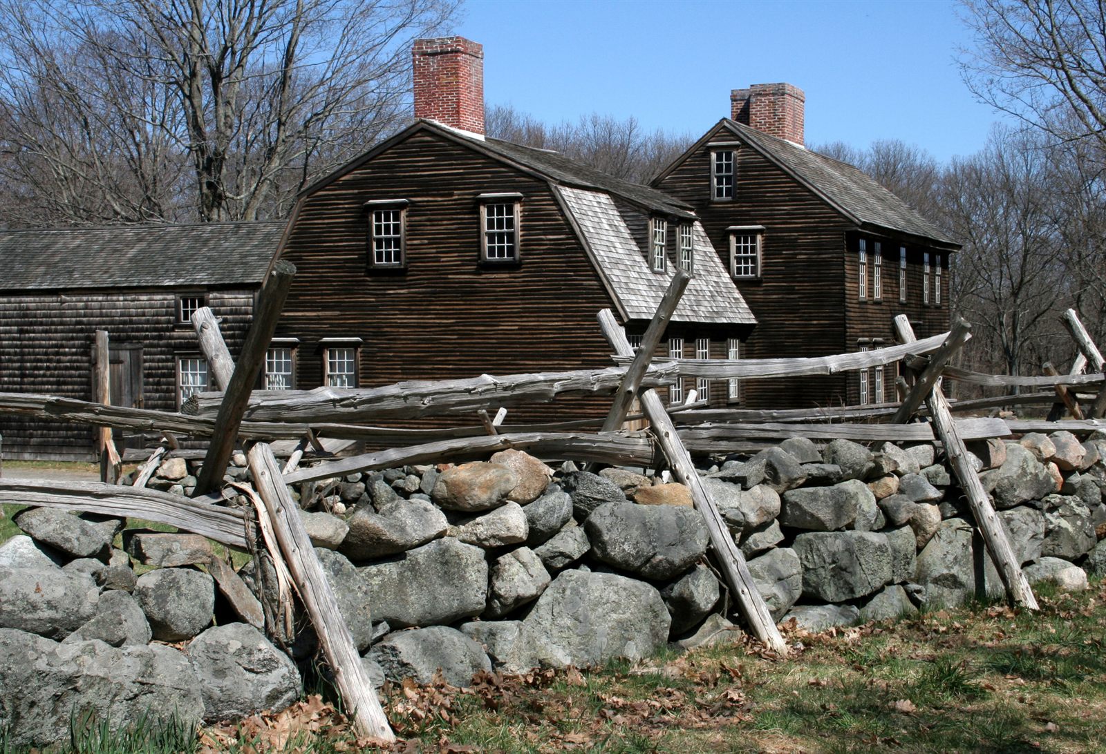 Weathered colonial-style wooden houses behind a fieldstone wall and split-rail fence